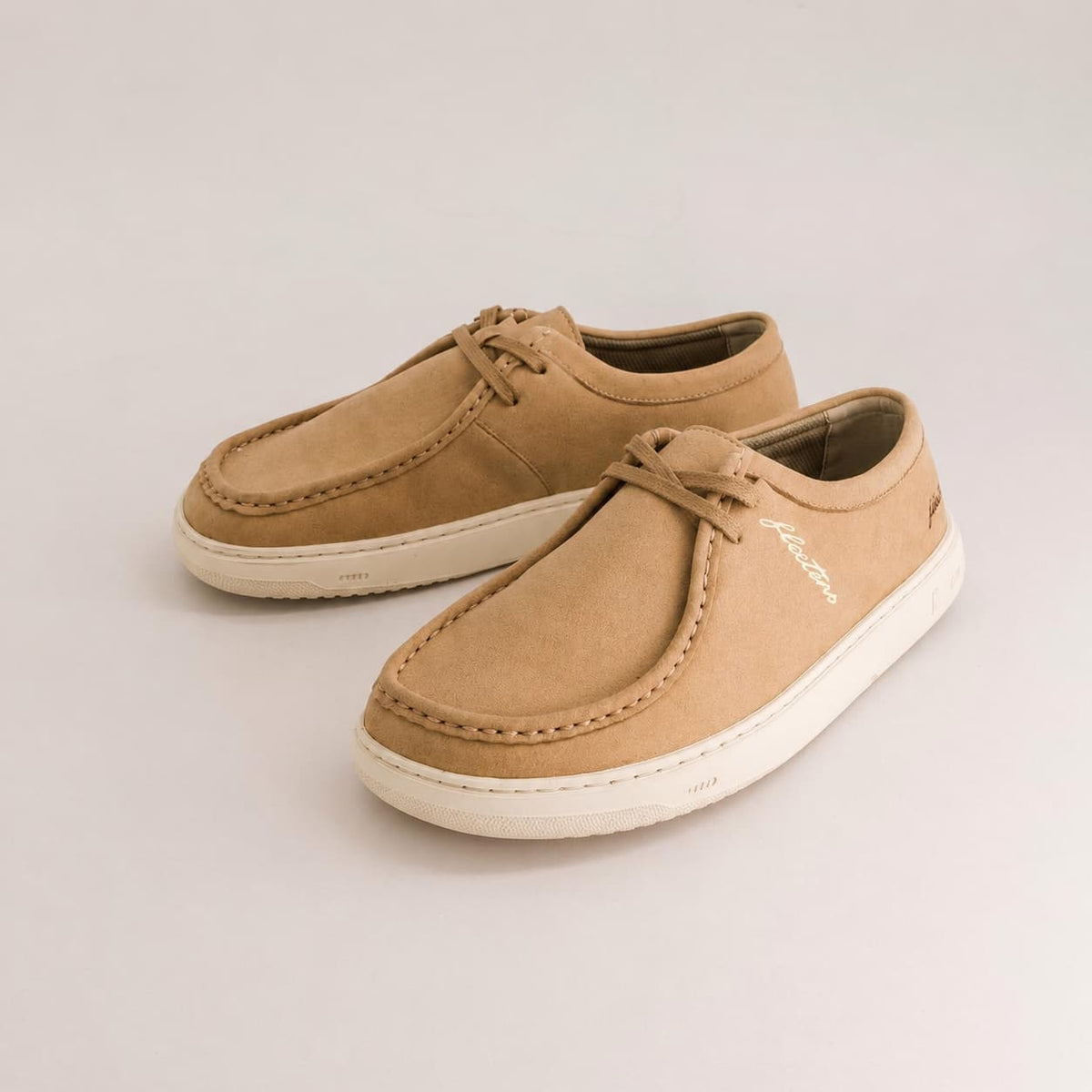 A pair of tan suede barefoot derby shoes with fringe details and white soles, photographed on a clean light gray background.