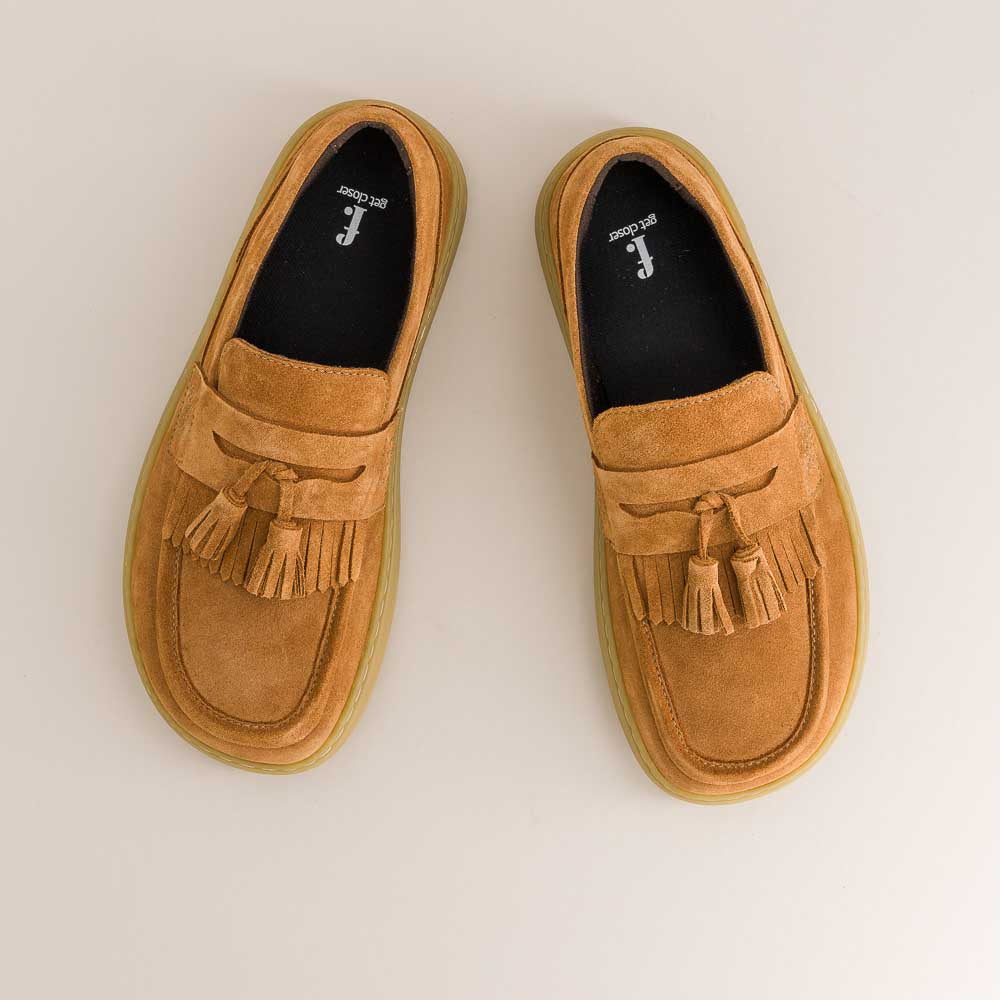 Pair of tan barefoot loafers with tassels on a light beige background