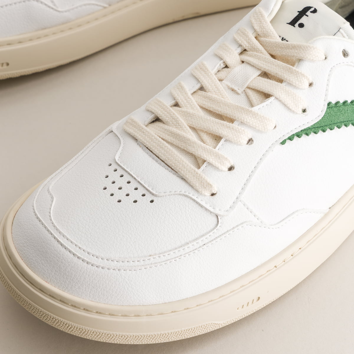 Close-up of a white barefoot sneaker with beige laces and green accents on a light background