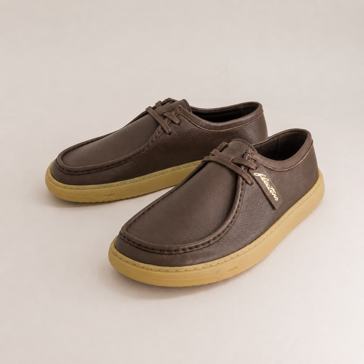 A pair of dark brown leather lace-up shoes with tan soles, displayed on a light gray background. A small gold brand tag is attached to the laces.