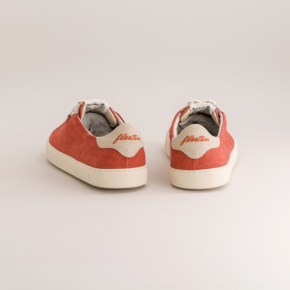 A pair of low-top barefoot sneakers shown from the back, featuring coral red suede uppers, cream leather heel tabs with the brand name 'Fleeters' embroidered in red, and off-white rubber soles, set against a plain light gray background.