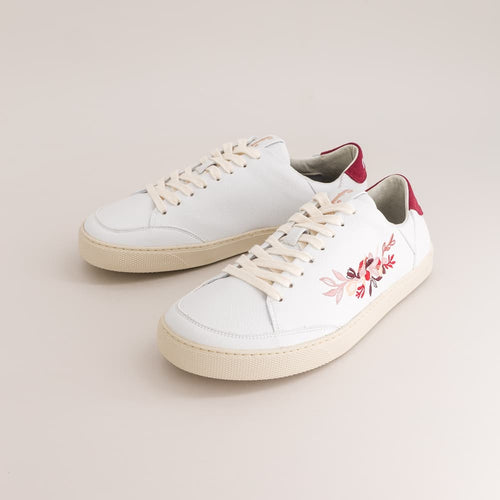 A pair of white leather barefoot sneakers with cream-colored soles and laces, featuring a pink and red floral embroidery on the side and a red heel tab, set against a solid light beige background.