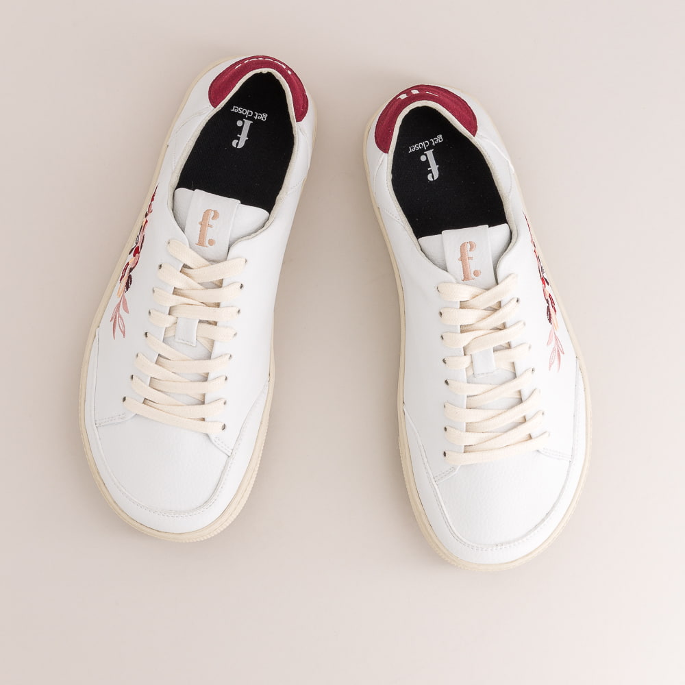 A pair of white leather barefoot sneakers with cream soles, featuring a floral embroidery on the side in pink, red, and green, and a burgundy heel tab with white lettering.