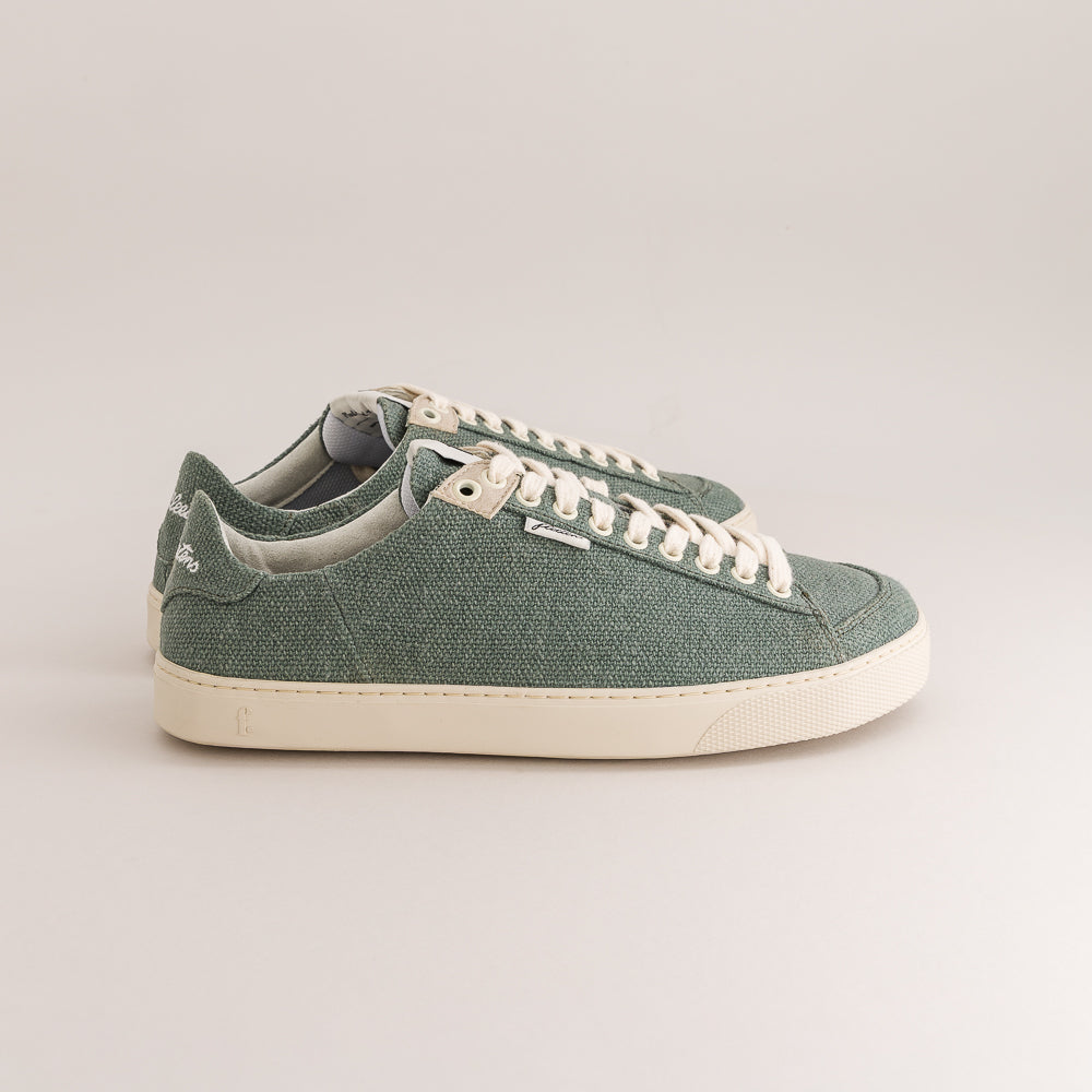 A pair of low-top wool sneakers in a muted sage green color, featuring off-white laces and a thick off-white rubber sole, photographed against a neutral light gray background.