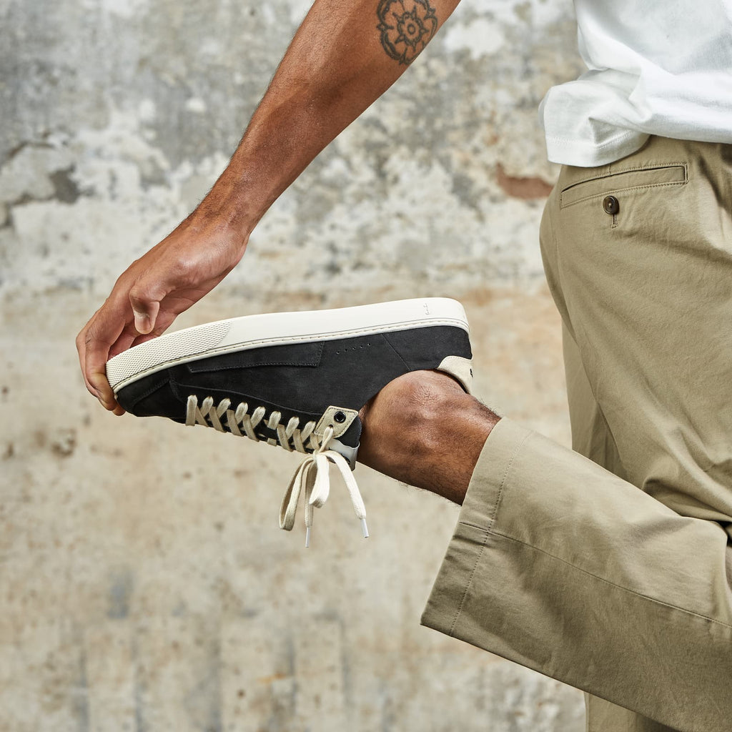 A person wearing a black barefoot sneaker with white laces and a white rubber sole, shown from the side as they bend their leg. The person is also wearing khaki pants and a white t-shirt, set against a textured, weathered concrete wall.