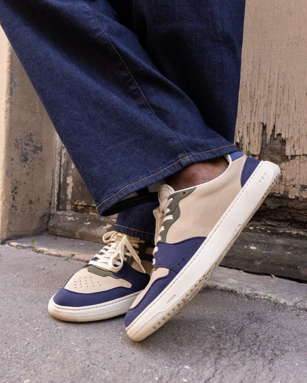 A person wearing navy and beige barefoot sneakers with dark green accents, paired with dark blue jeans, standing on a concrete surface.