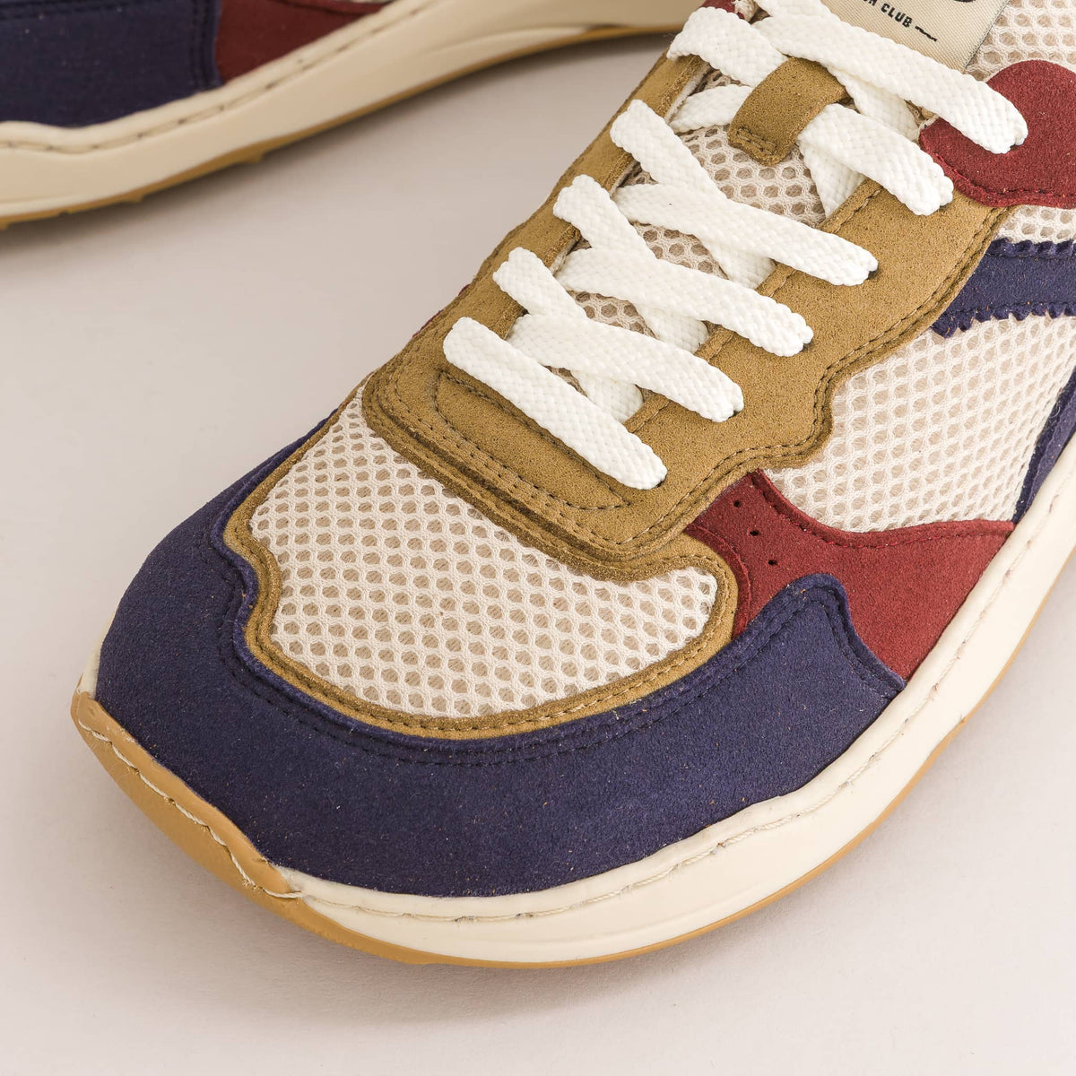A close-up of a multi-colored retro barefoot sneaker featuring navy blue suede, beige mesh, tan suede overlays, burgundy panels, and white laces.