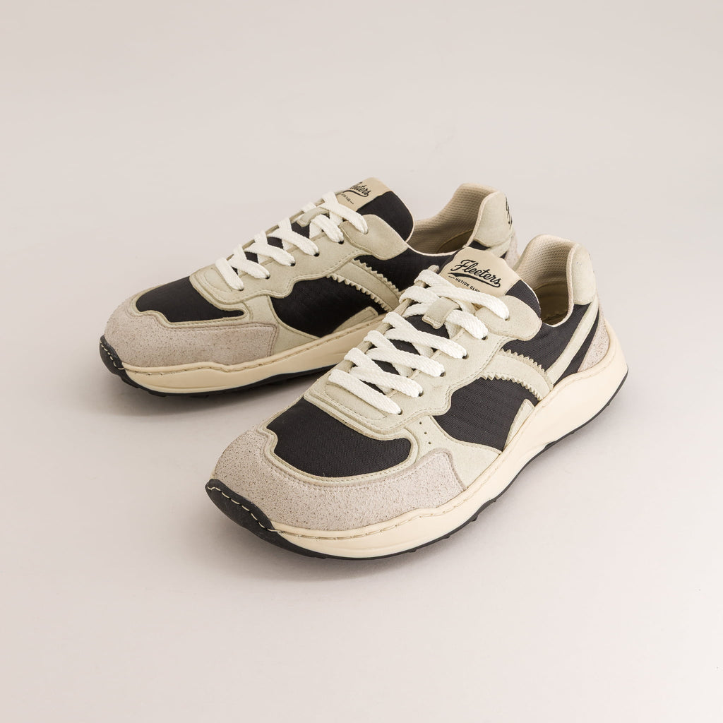 A pair of retro-style low-top barefoot sneakers in beige, black, and off-white, featuring white laces and a chunky cream midsole, photographed on a light beige studio background.