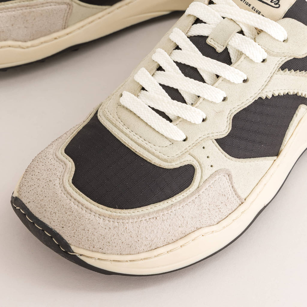 A close-up of a low-top barefoot sneaker showing the toe box, white laces, and layered panels in off-white suede and black mesh.