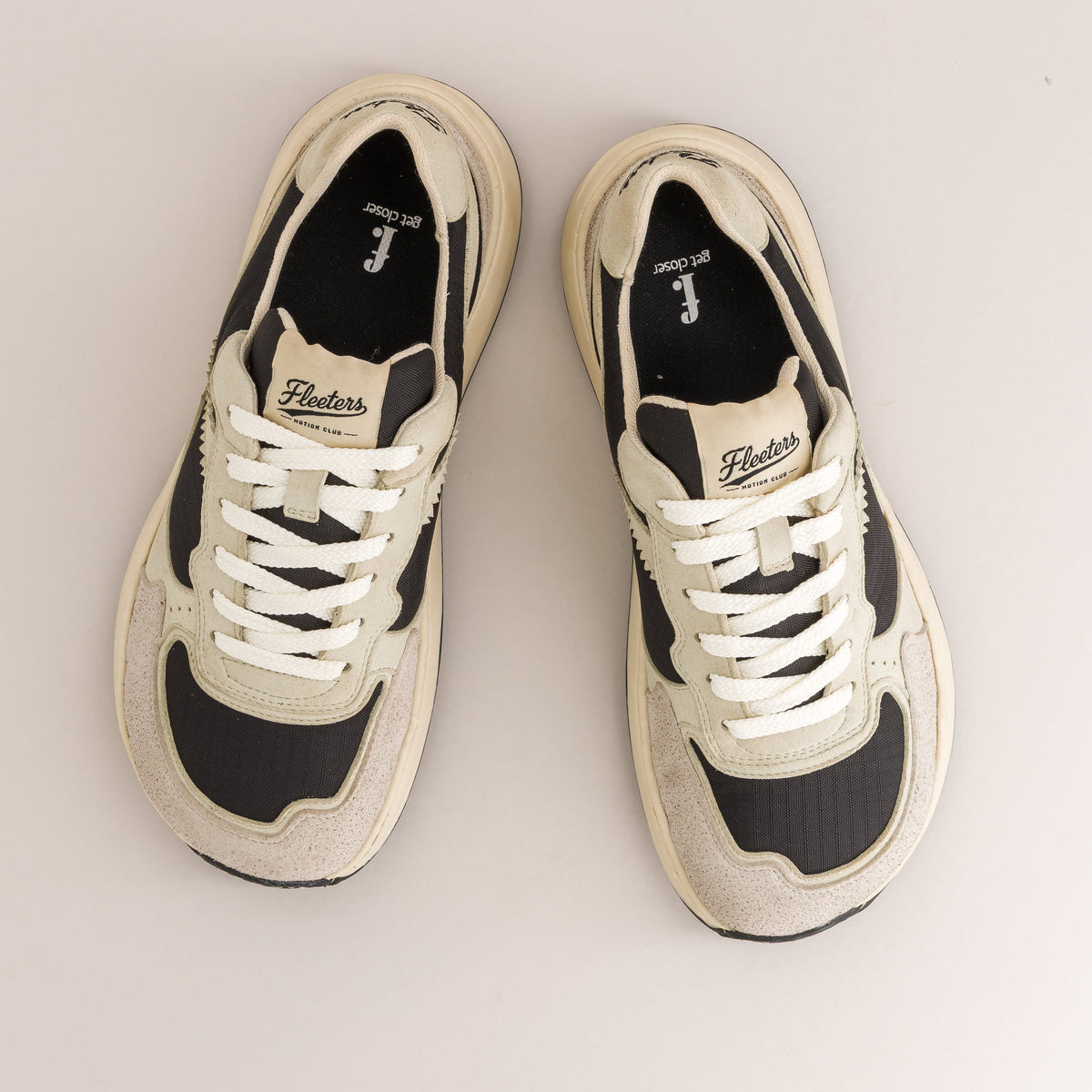 A pair of low-top barefoot sneakers shown from a top-down view on a light beige background. The shoes feature cream-colored suede and mesh panels, black synthetic overlays, and white laces.