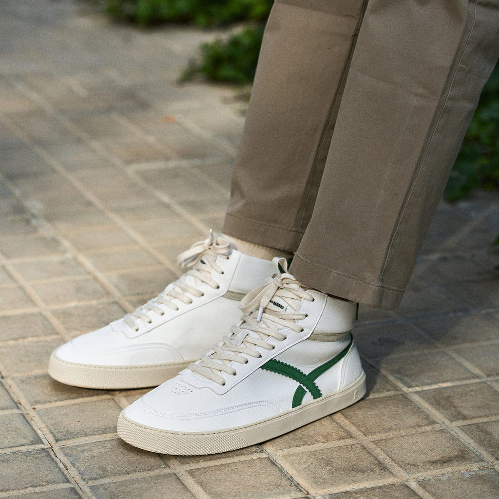White high-top barefoot sneakers with green accents worn by a person on a paved surface.