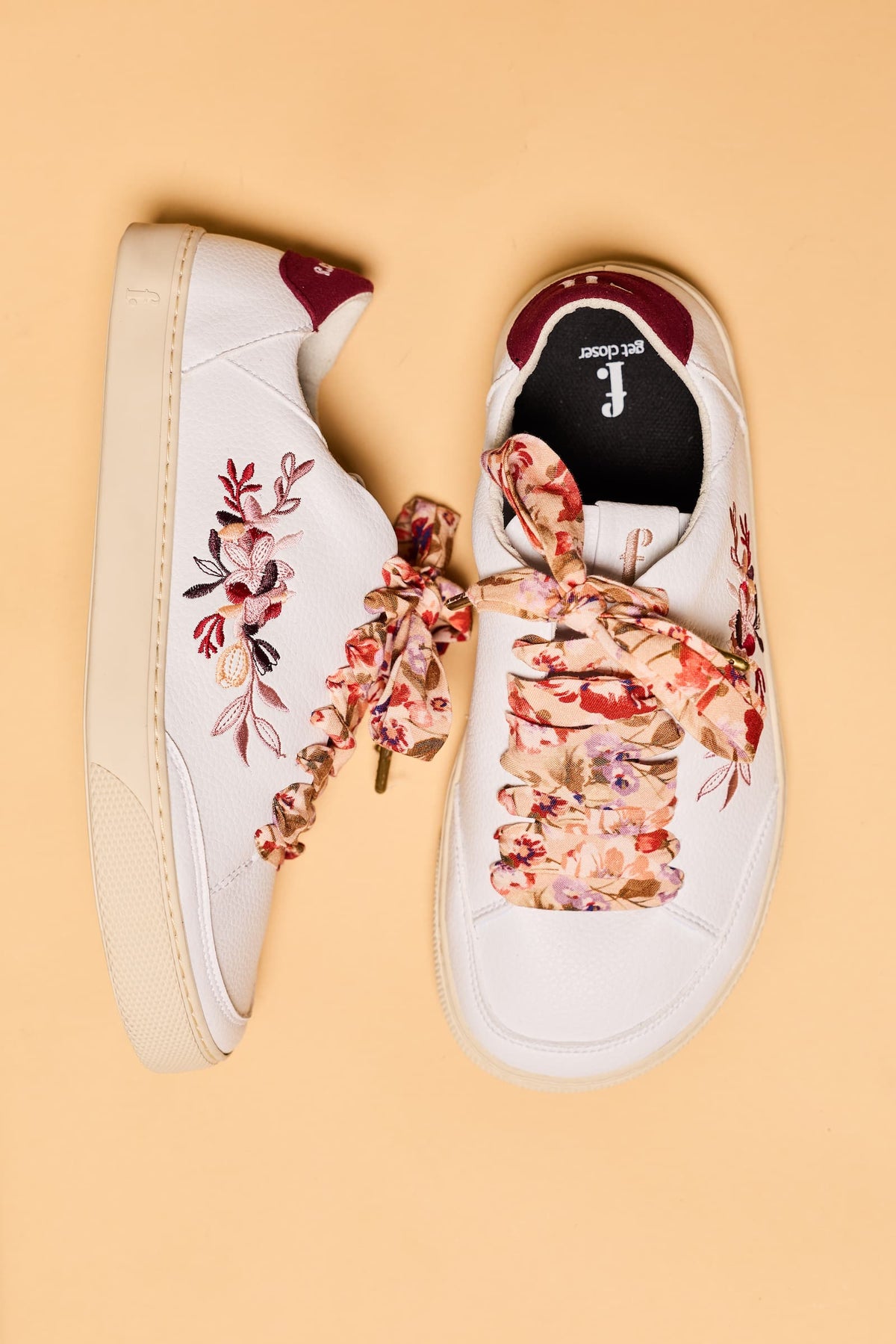 A pair of white low-top barefoot sneakers with floral embroidery and matching floral ribbon laces, displayed on a solid beige background.