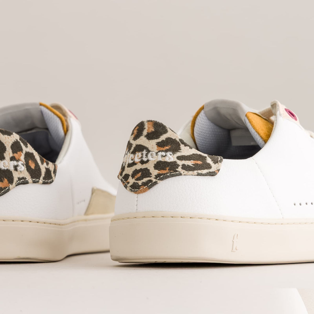 White leather barefoot sneakers with leopard-print heel tabs and cream soles, photographed on a clean light beige studio background.