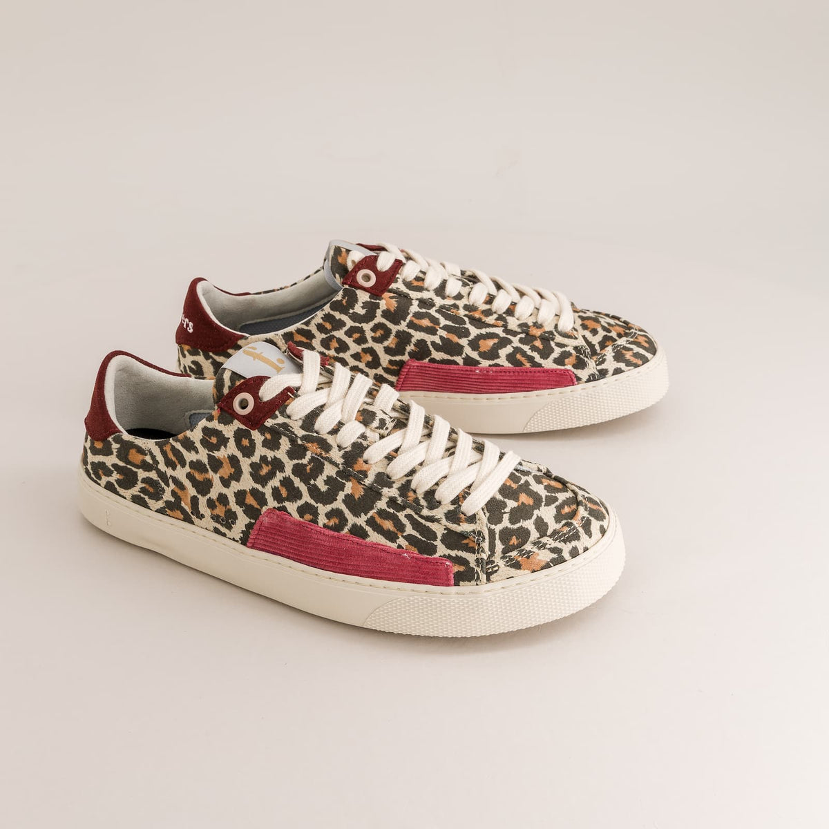 A pair of low-top barefoot sneakers featuring a beige and brown leopard print pattern, white laces, and burgundy accents on the heel tab, side stripe, and tongue label, set against a plain light gray background.