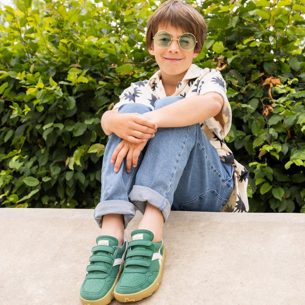 Un jeune garçon portant des lunettes de soleil rondes teintées de vert, une chemise à motifs blanche à manches courtes, un jean bleu et des baskets vertes, assis sur un rebord en béton devant une haie verte.