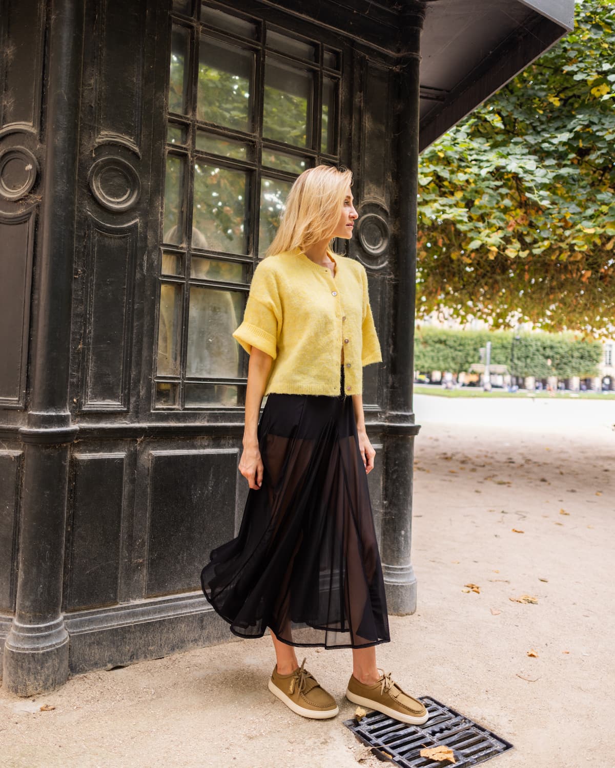 A woman stands outdoors next to a dark building, wearing a short-sleeve yellow knit top, a long black pleated midi skirt, and barefoot derby shoes.