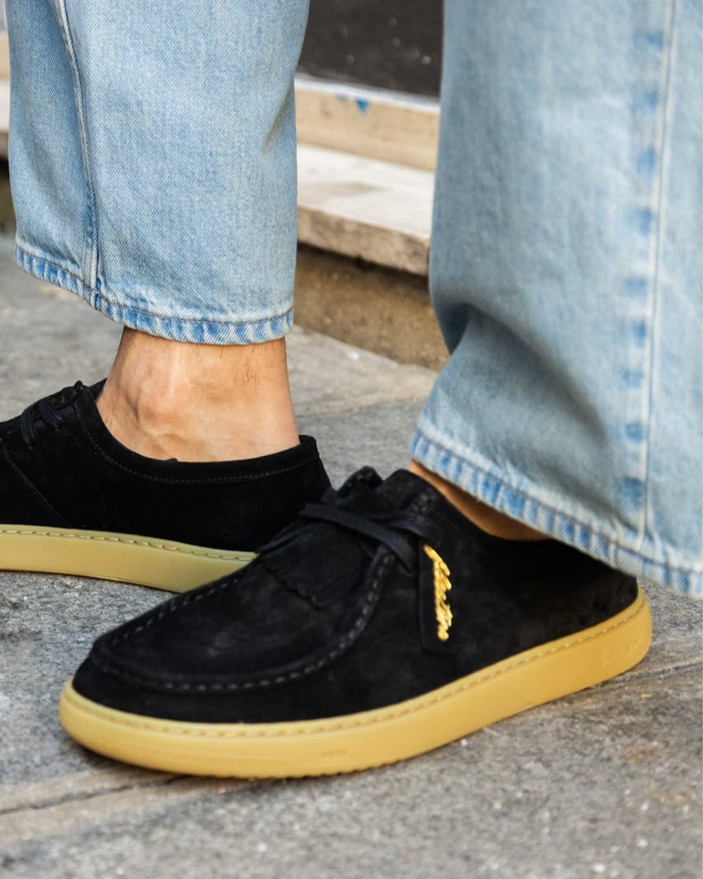 Black sneakers with yellow soles worn with light blue jeans on a concrete surface.