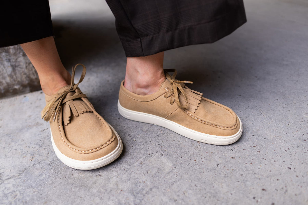 A close-up of a person wearing tan suede barefoot derby shoes with white soles, standing on a gray concrete floor.