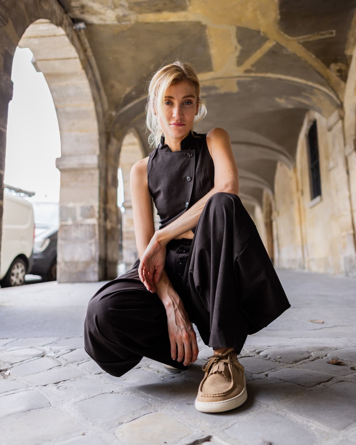 A woman crouches in a stone arcade wearing a black sleeveless top and wide-leg pants with beige barefoot derby shoes.