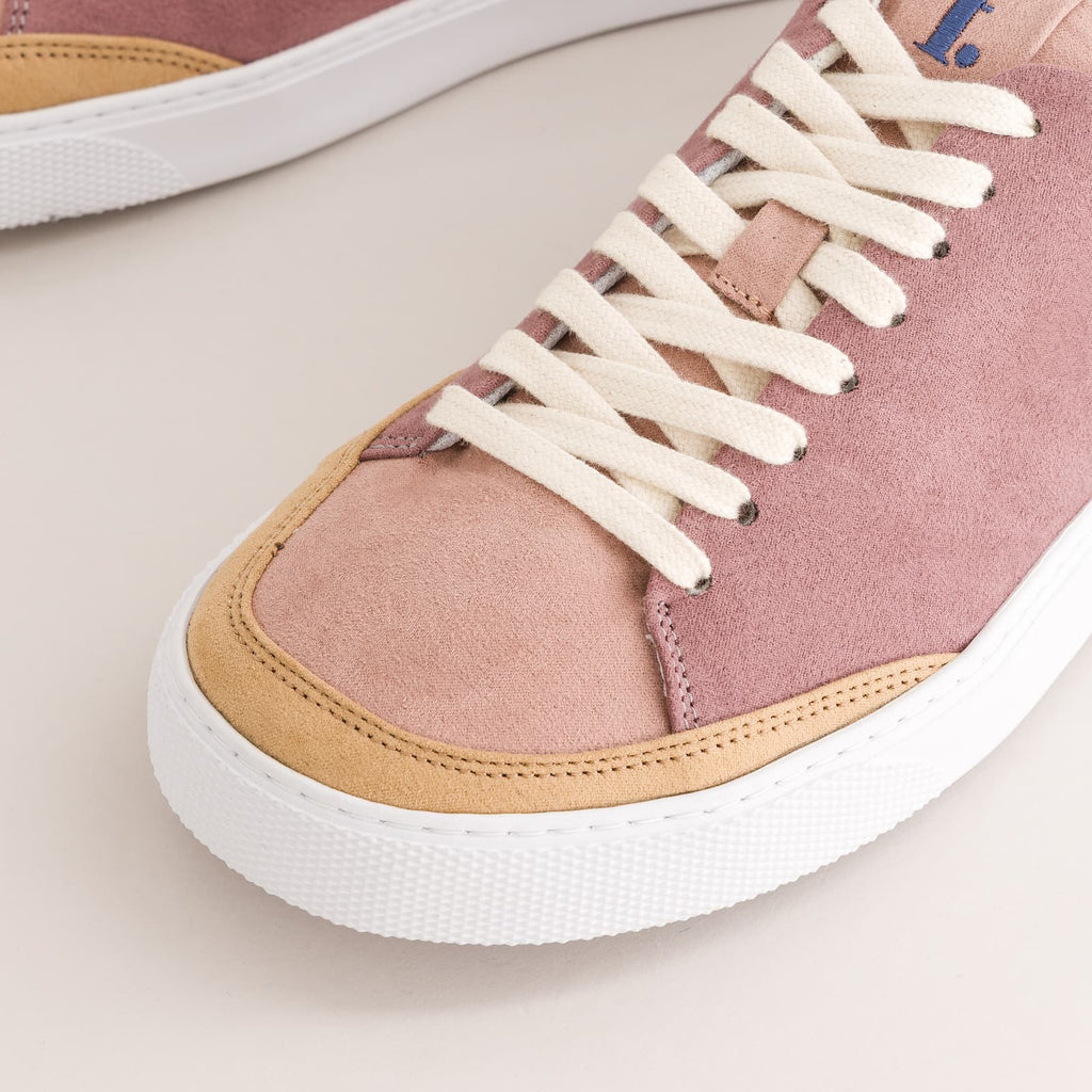 A close-up of a low-top barefoot sneaker in dusty pink suede with cream laces, tan suede trim, and a thick white rubber sole, photographed on a light beige background.